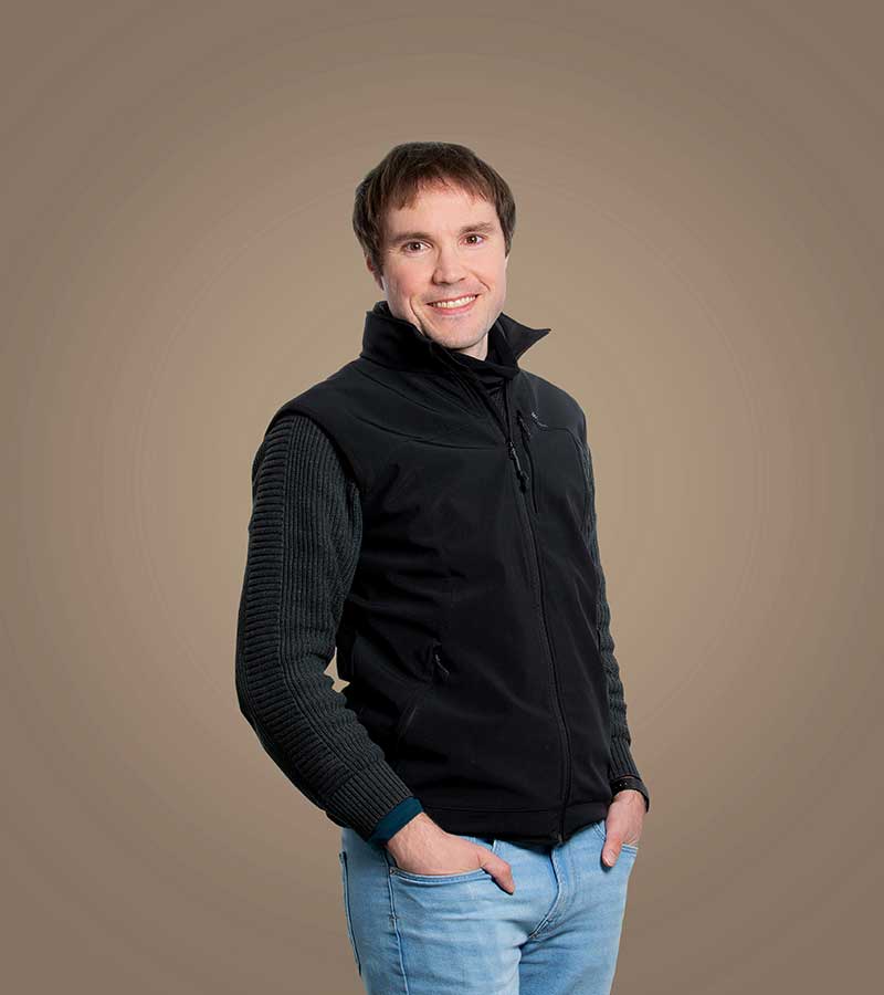 Baumberge Verein Porträt eines lächelnden Mannes mit kurzen braunen Haaren, der eine schwarze Jacke mit Reißverschluss trägt, vor einem grünen Hintergrund.