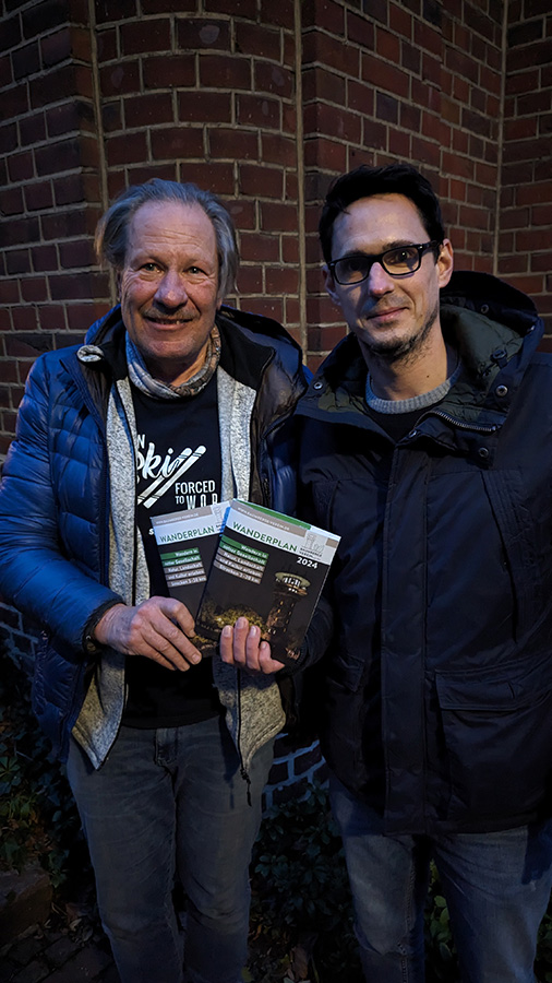 Baumberge Verein Zwei Männer stehen vor einem Backsteingebäude und halten Bücher in der Hand.